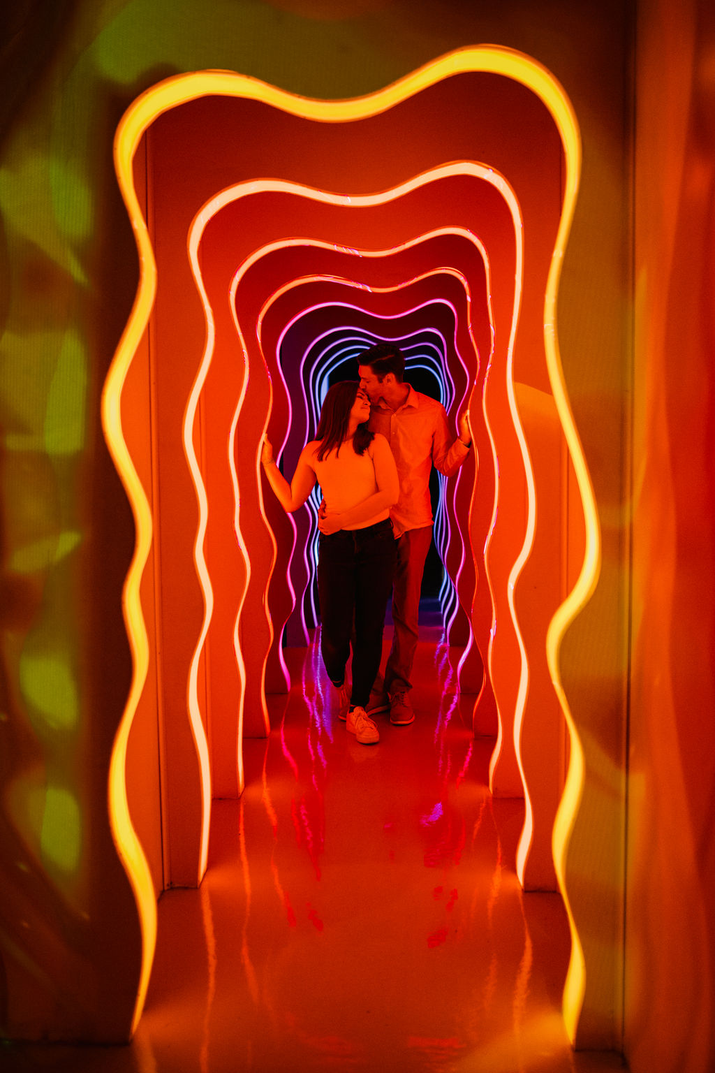 Liz and Justin walk through a neon red and yellow tunnel made of waves.