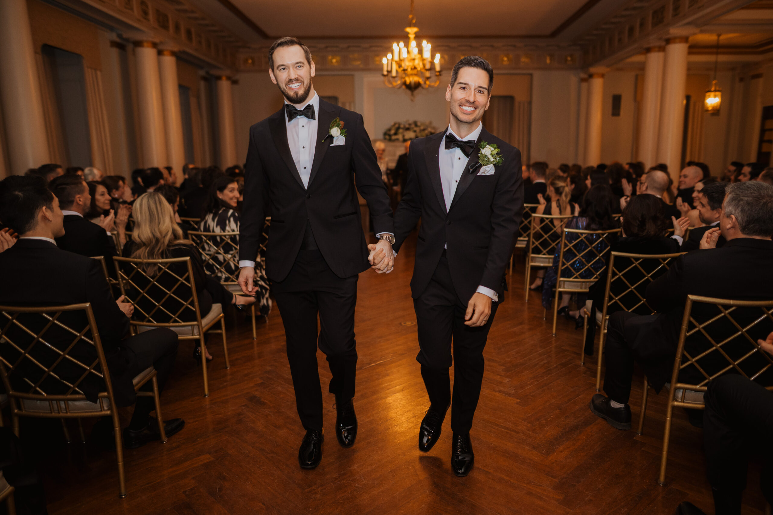 A gay couple in tuxedo attire walk down the aisle of their formal wedding at the Cincinnati Club.